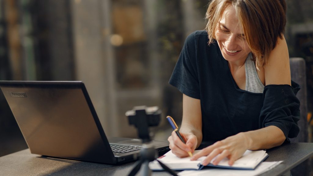 femme heureuse et concentrée à travailler qui écrit sur un carnet avec son ordinateur à coté. 
